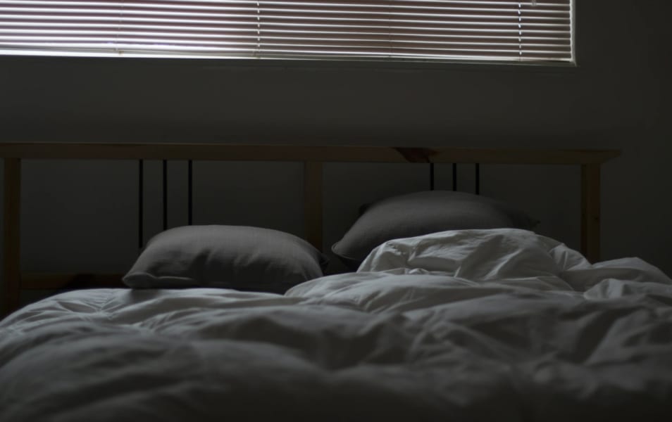 Dark bedroom with light coming through the blinds and an unmade but cozy bed