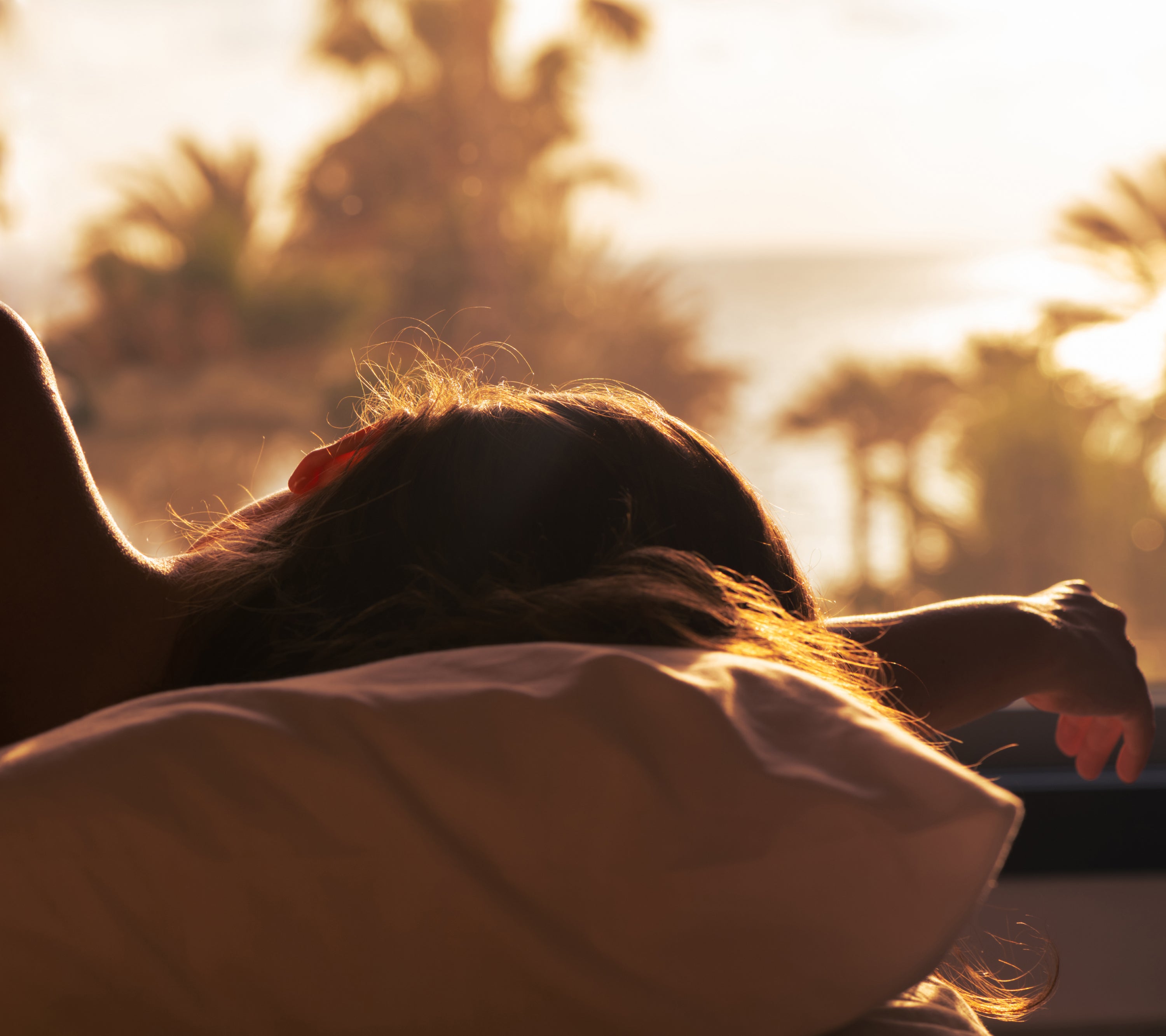 Silhouette of a woman sleeping on a pillow with a sunrise behind her with palm trees and ocean.