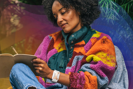 Women sitting writing in journal in front of tree wearing coloring fleece jacket with an Apollo wearable on her left wrist