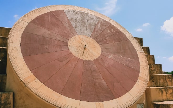 A circular solar clock made of stone. There are two circles that are different tines of red in front of a stone stair case with a blue sky.