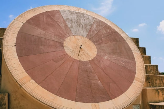 A circular solar clock made of stone. There are two circles that are different tines of red in front of a stone stair case with a blue sky.
