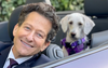 Man in suit with curly hair in convertible with dog in purple harness behind him